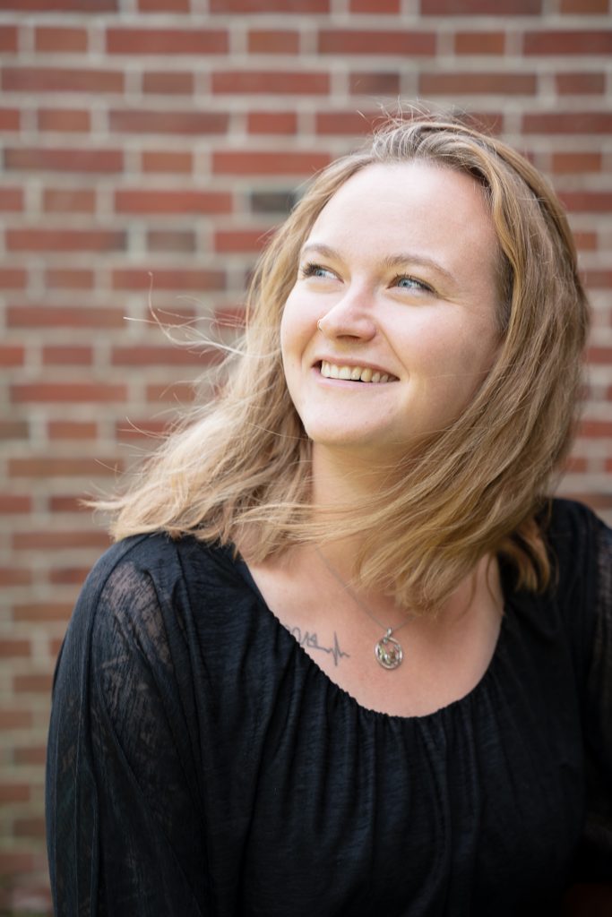 Juliane Kau sitzt vor einer Ziegelmauer. Sie schaut nach links oben, lächelt und ihre Haare sind vom Winde verweht.
