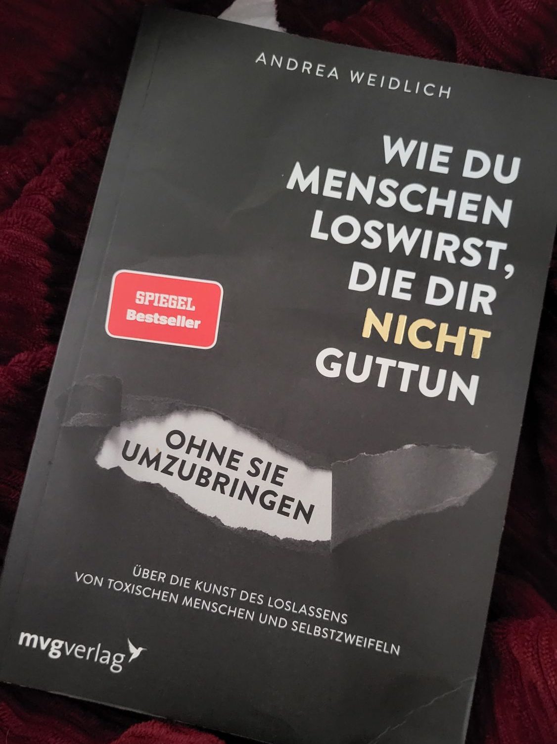 Das Buch dem Titel "Wie du Menschen loswirst, die dir nicht guttun" und Untertitel "Ohne sie umzubringen" von der Autorin Andrea Weidlich, ist mit einem orangefarbenen Aufkleber "SPIEGEL Bestseller" versehen. Es liegt auf einer bordeauxfarbenen Tagesdecke auf dem Schoß der Bloggerin Juliane.