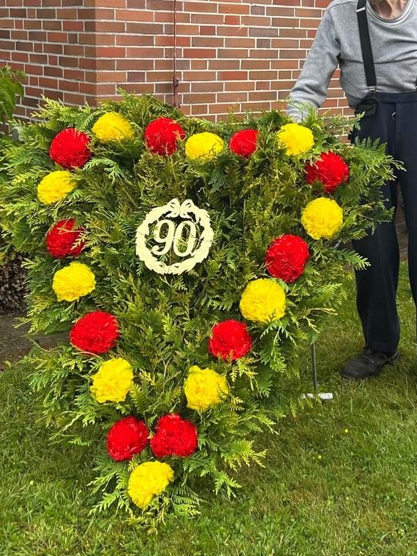 Ein Herbogen ist zu sehen, der mit grünem Lebensbaum, roten und gelben Rosen aus Papier und eine goldenen 90 verziert ist. Ein älterer Mann steht dahinter. Sein Kopf und Gesicht sind nicht zu sehen.