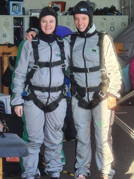 Die Bloggerin Juliane steht mit ihrer Freundin Tina Seite an Seite. Sie sind mit einem Anzug, einer Haube und einem Geschirr ausgestattet, um mit ihren Tandempartner Fallschirm zu springen.