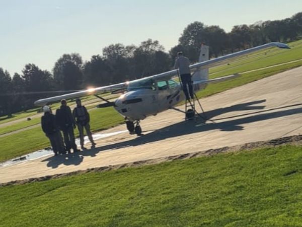 Zu sehen ist eine kleine Propeller Maschine um die vier Menschen herum stehen. Der Pilot tankt das Flugzeug über die Tragflächen auf und gleich geht an den Start, um die Fallschirmspringer in luftige Höhen zu bringen.