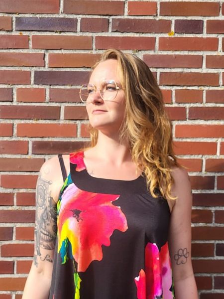 Bloggerin Juliane Kau steht vor einer Ziegelsteinwand. Sie trägt eine Brille, ein buntes Kleid und ihre Haare offen. Ihr Gesicht ist der Sonne zugewandt. 