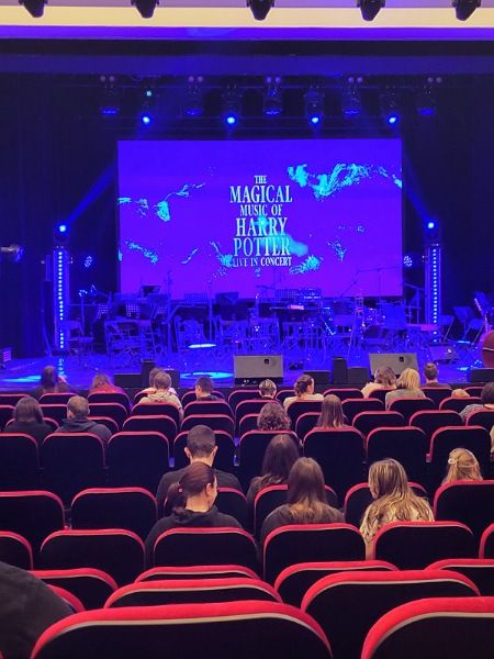 Es ist der Konzertsaal zu sehen. Auf der Bühne wir auf einer Leinwand der Titel der Veranstaltung "Harry Potter in concert" eingeblendet. Der Saal füllt sich allmählich mit Menschen, die auf den roten Polsterstühlen Platz nehmen.