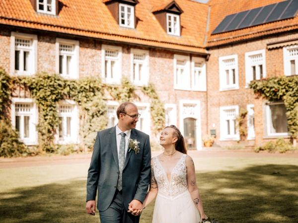 Die Bloggerin Juliane Kau geht mit ihrem Ehemann über den Rasen vor der Burg Berum. Dort hat sich das Brautpaar am 19. September 2025 trauen lassen. Beide strahlen vor Glück.