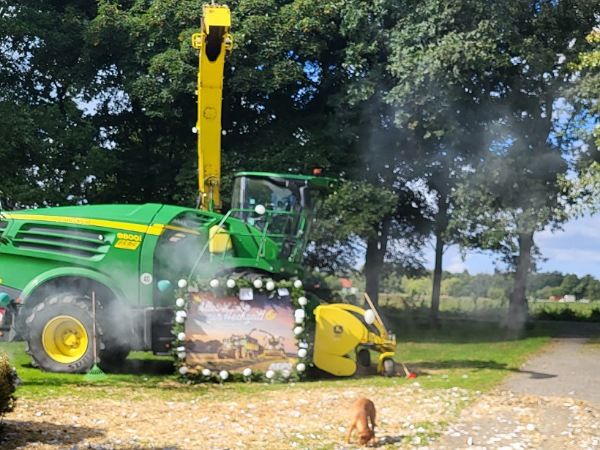 Zu sehen ist ein Feldhäcksler der Marke John Deere. Davor liegt ganz viel gehäckseltes Stroh und Hündin Pina schnüffelt darin. An den Feldhäcksler ist ein großer Bogen gelehnt mit den besten Wünschen zur Hochzeit.
