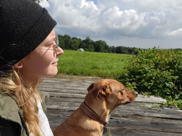 Die Bloggerin Juliane Kau sitzt mit Hündin Pina auf einer Holzbrücke. Sie trägt eine Brille und eine Mütze - also die Bloggerin, nicht der Hund. Beide schauen in eine Richtung aufs Wasser. 
